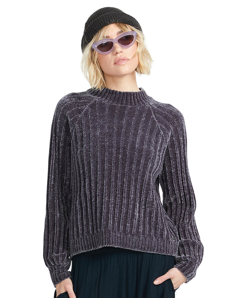 Female model wearing a ribbed velvet-textured dark sweater, black beanie, and purple sunglasses, posing against a white backdrop.