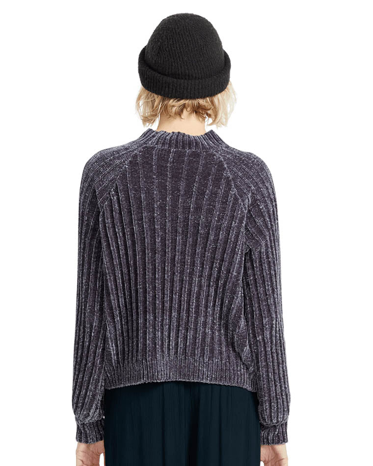 Back view of a female model wearing a ribbed dark velvet-textured sweater and a black beanie, standing against a white backdrop.