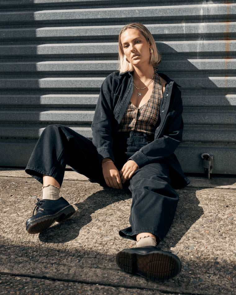 Model sitting against a metal wall wearing a black oversized denim jacket, plaid crop top, wide-leg pants, and black lace-up shoes.