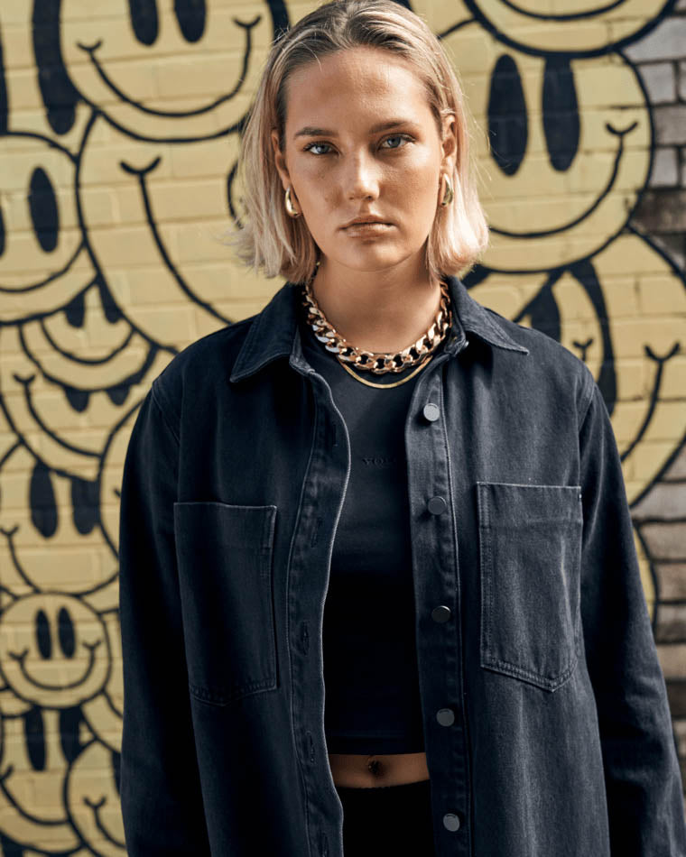 Model in a black oversized denim jacket and crop top, wearing chunky gold jewelry, standing against a yellow smiley face mural.