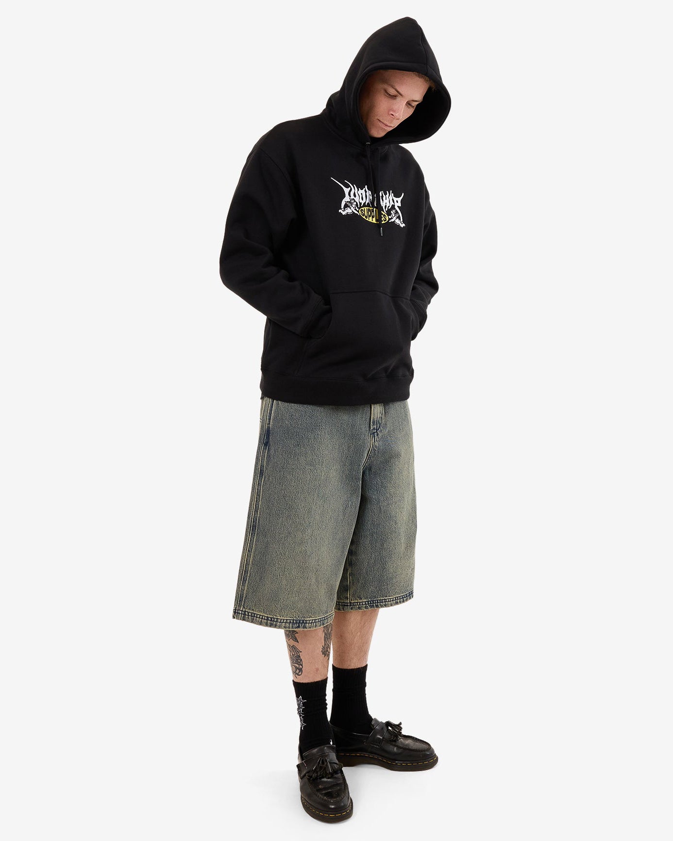 Male model wearing a black graphic hoodie with the hood up, paired with oversized denim shorts, black socks, and loafers.