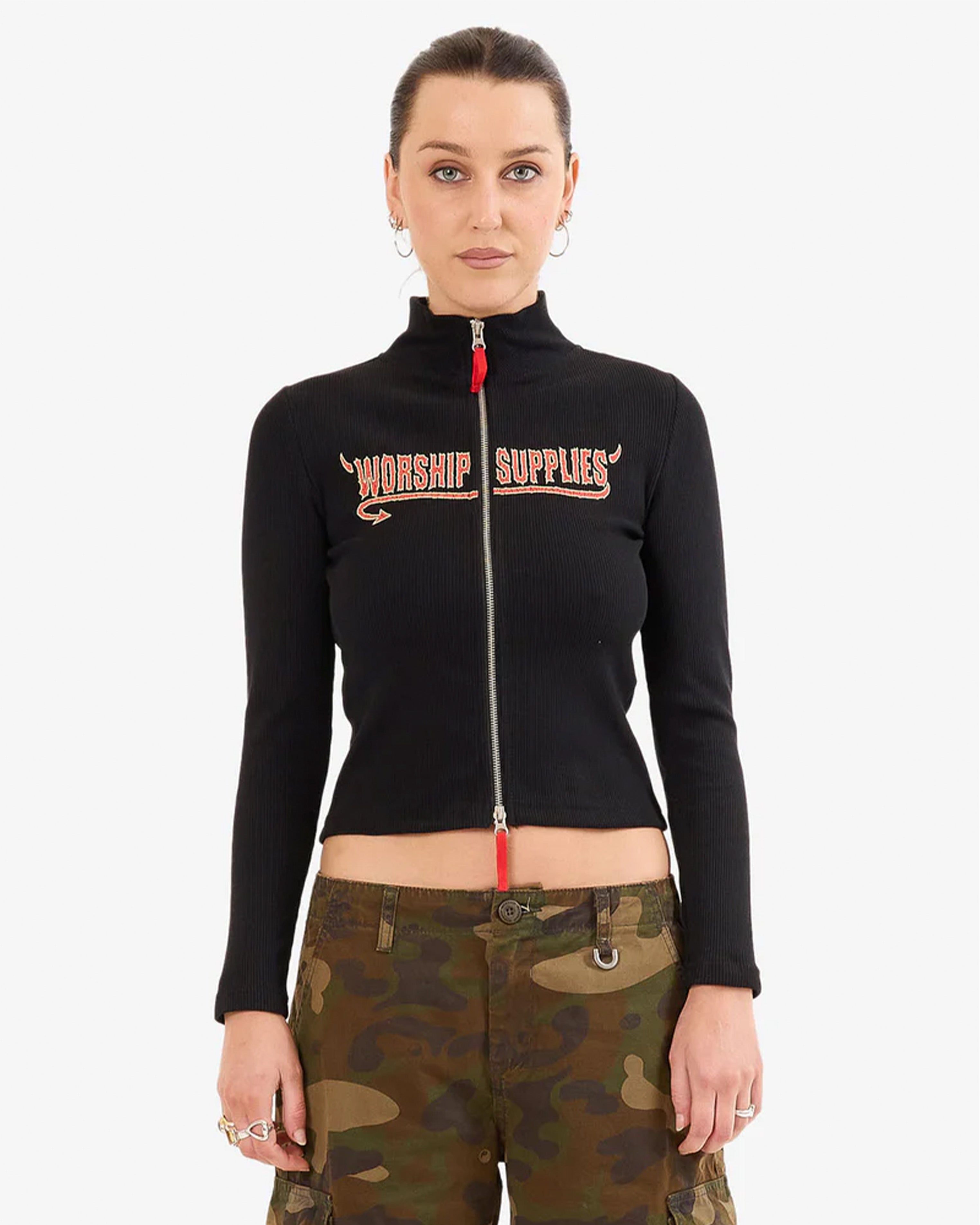 Woman front view wearing a fitted black zip-up top with “WORSHIP SUPPLIES” text, paired with camo cargo pants and hoop earrings.
