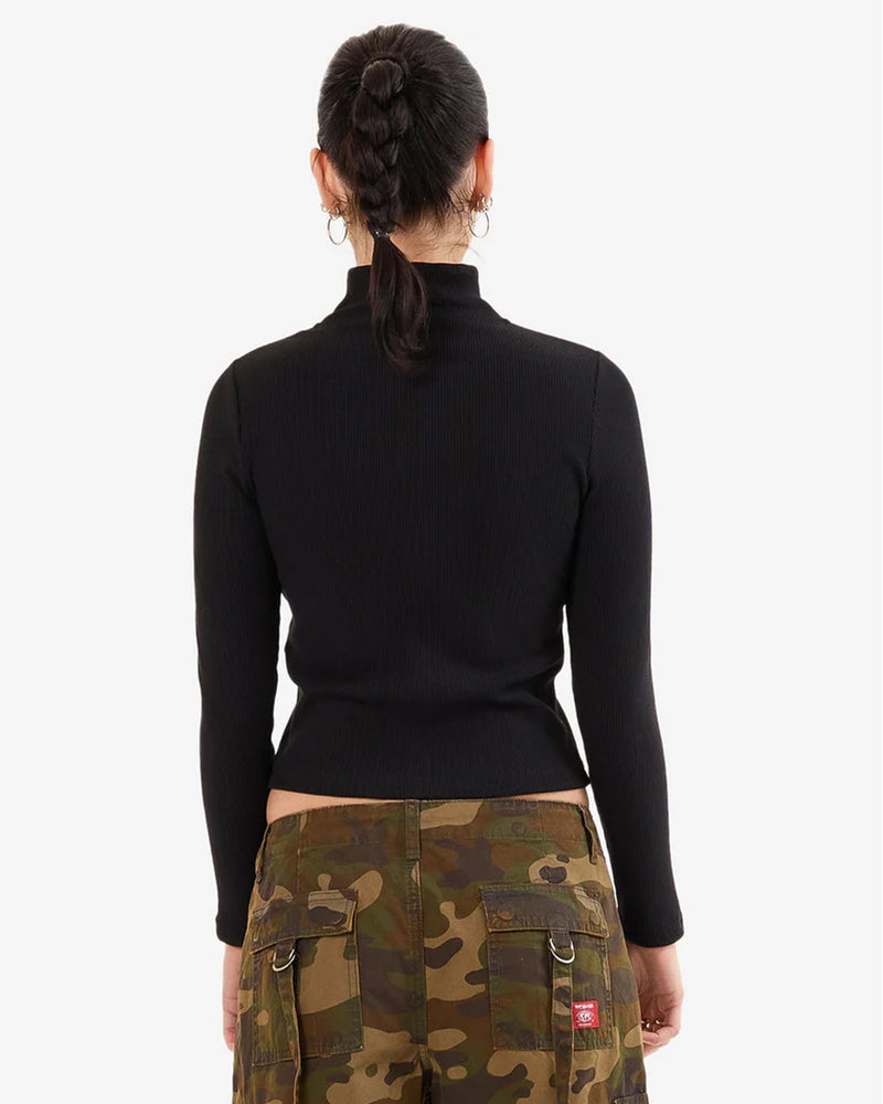 Woman back view wearing a fitted black long sleeve zip-up top and camo cargo pants, with hoop earrings and a braided ponytail.
