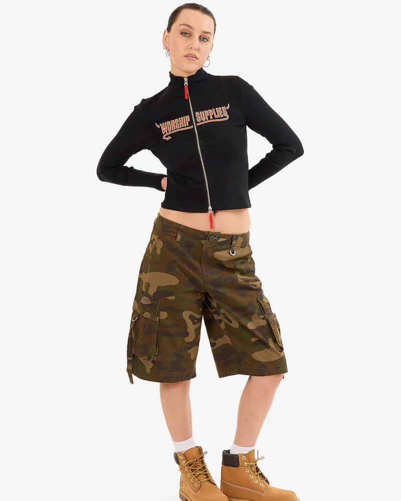 Woman front view wearing a fitted black “WORSHIP SUPPLIES” zip-up top, camo knee-length cargo shorts, and tan boots.
