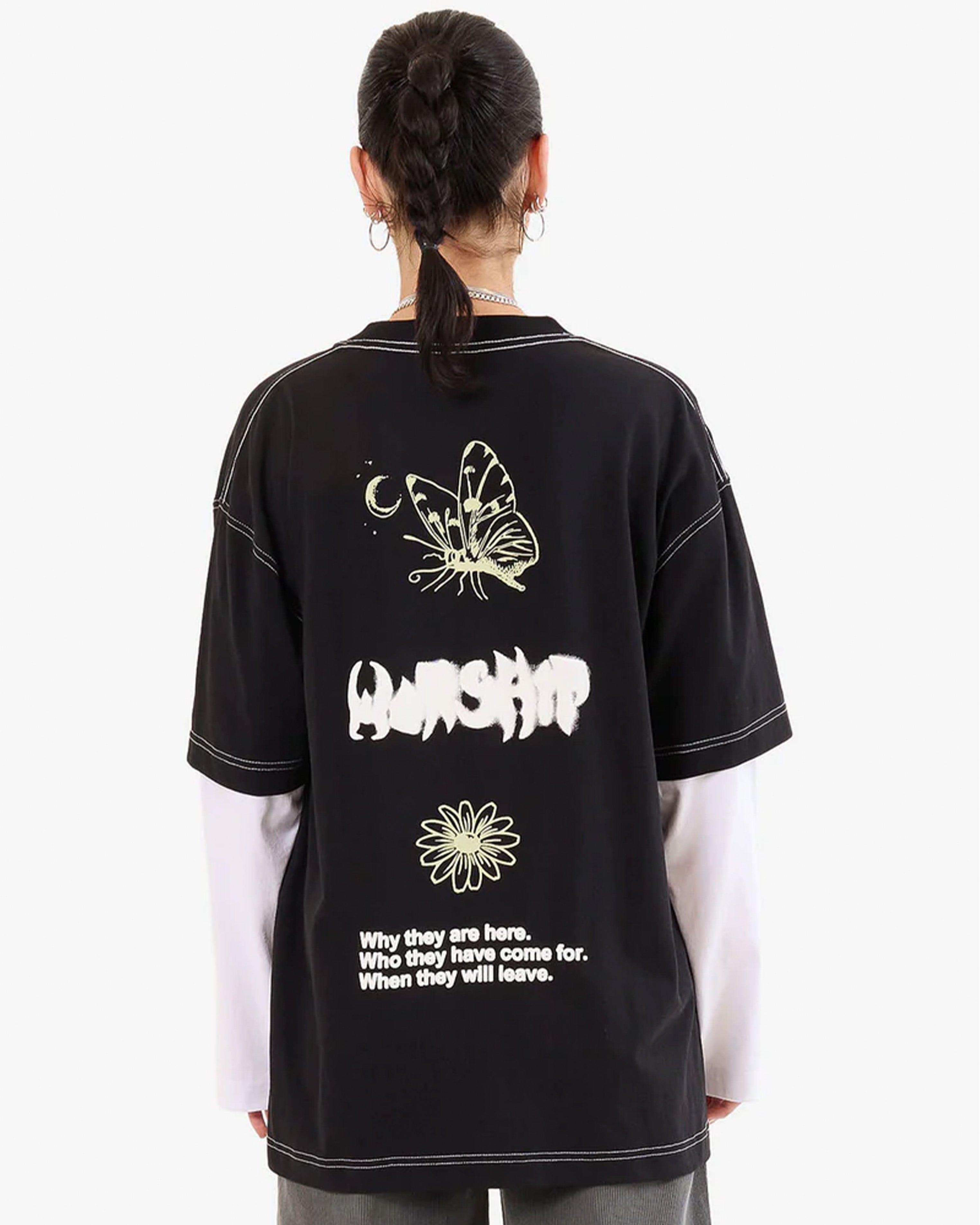 Woman back view wearing a black oversized graphic tee with white text, butterfly and flower prints, layered over long white sleeves.
