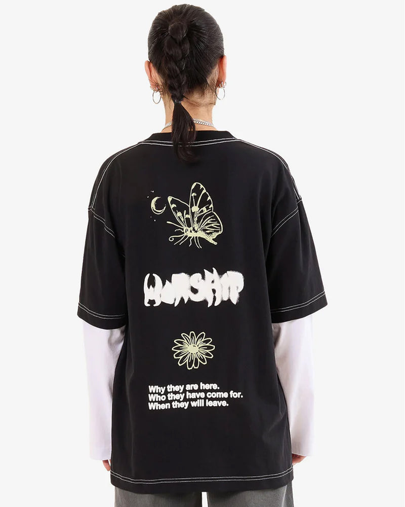 Woman back view wearing a black oversized graphic tee with white text, butterfly and flower prints, layered over long white sleeves.
