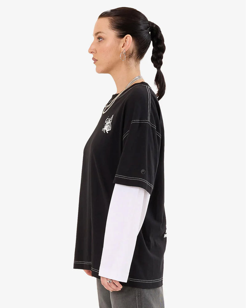 Woman left-side view wearing a black oversized graphic tee layered over long white sleeves, with hoop earrings and a braided ponytail.
