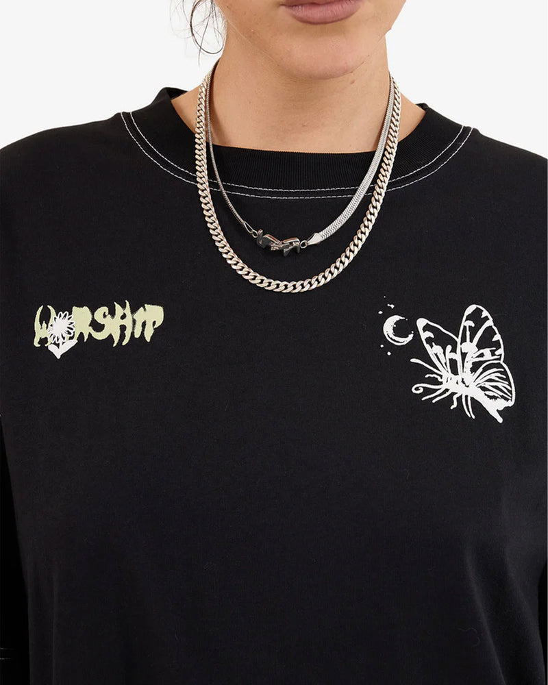 Close-up front view of a woman wearing a black graphic tee featuring a small "WORSHIP" logo and a butterfly with a crescent moon, worn with layered silver chains.