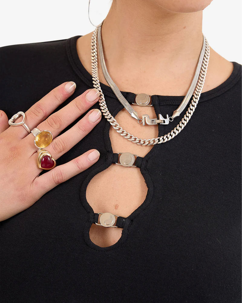 Close-up front view of a black long sleeve top featuring three circular metal clasp details along a front cut-out design, with layered silver necklaces and rings visible.