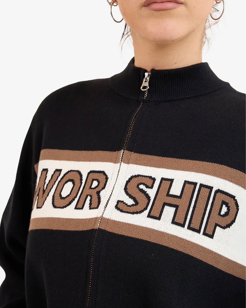 Close-up of a black zip-up jumper with “WORSHIP” text across a beige and white chest stripe, worn by a woman.