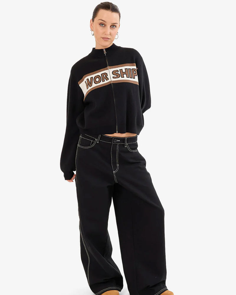 A full-body image of a woman wearing a black zip-up sweater with 'WORSHIP' text, wide-leg pants with contrast stitching, and hoop earrings.
