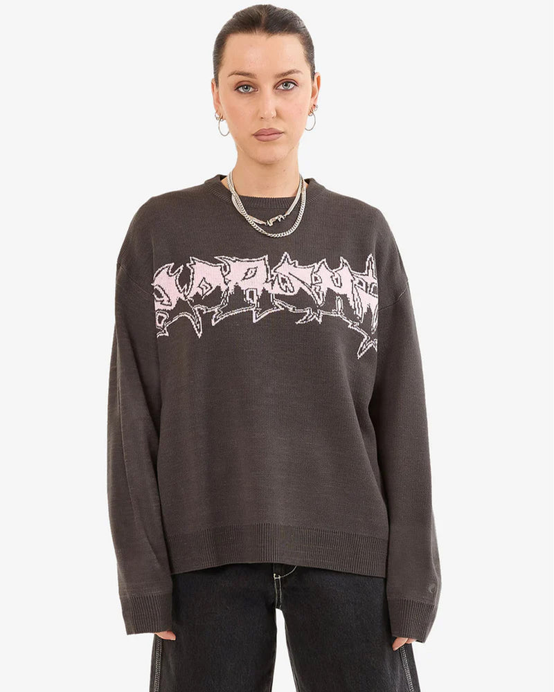 Woman front view wearing a dark oversized knit sweater with bold pink graffiti-style text, hoop earrings, and black wide-leg jeans.
